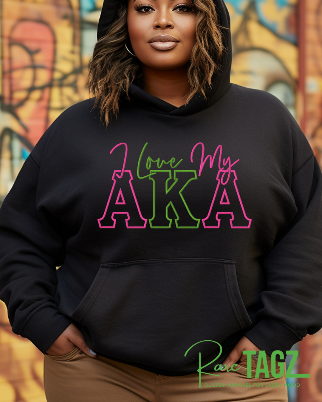 Person wearing a black hoodie with colorful text in front of a graffiti wall. Woman wearing a black hoodie with pink and green lettering that reads ‘I Love My AKA.’ The hoodie has a relaxed fit with a kangaroo pocket and drawstring hood. Background shows colorful graffiti art. Rare Tagz logo is in the corner.