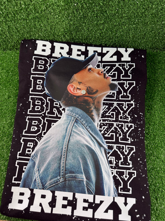 Chris Brown wearing a denim jacket with 'BREEZY' text on a green grass background. Black Breezy Bowl tour T-shirt with bold white lettering and Chris Brown graphic design. 