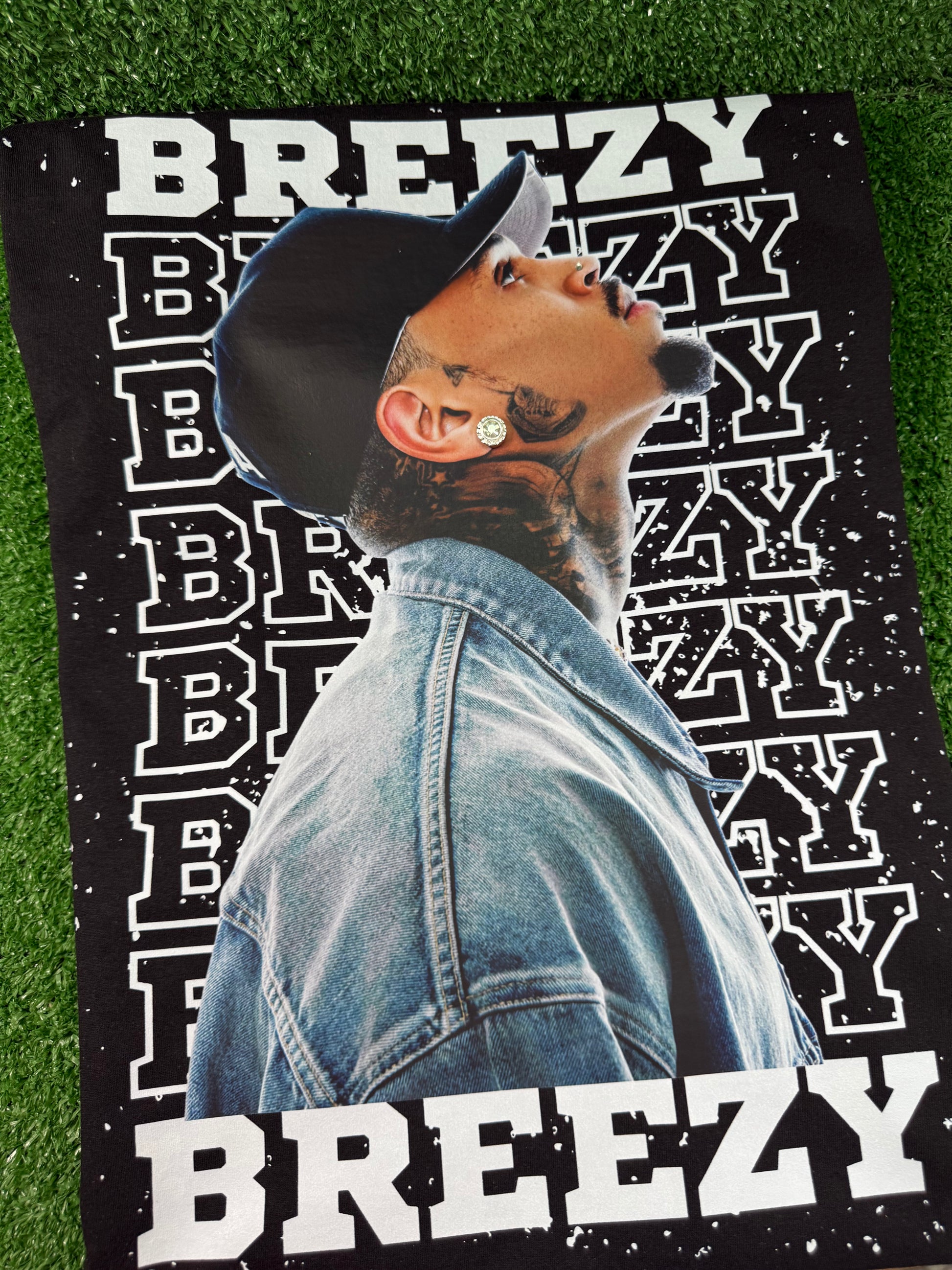 Chris Brown wearing a denim jacket with 'BREEZY' text on a green grass background. Black Breezy Bowl tour T-shirt with bold white lettering and Chris Brown graphic design. 