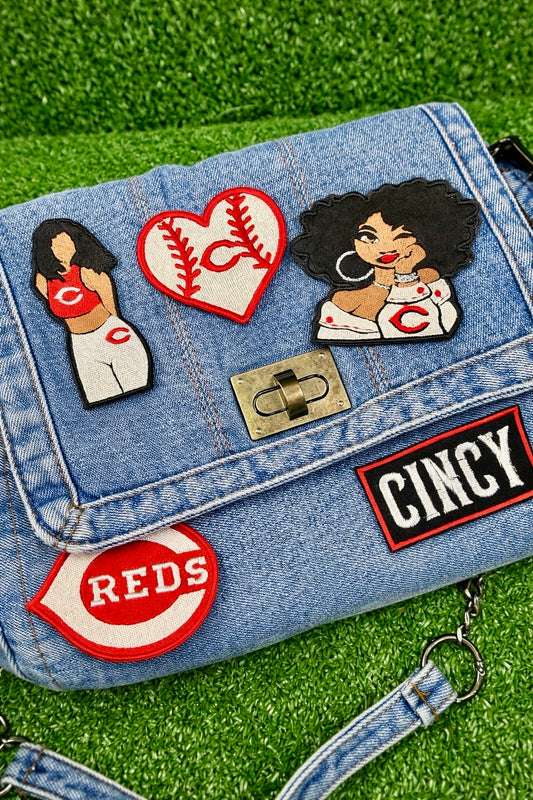 Denim bag with various patches including 'Cincy' and 'Reds' logos on a grass background
