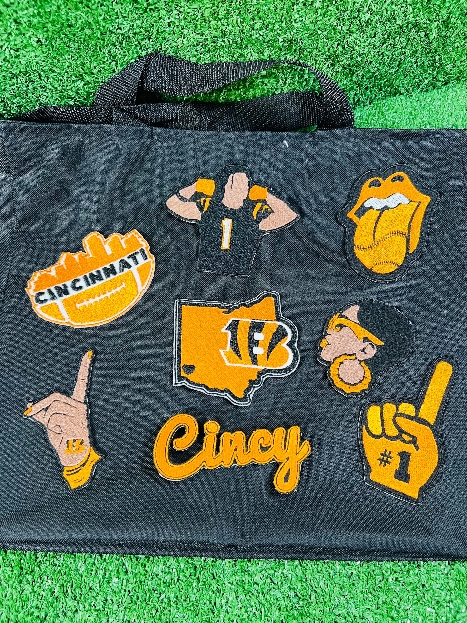 A black tote bag laid flat on green artificial turf, decorated with multiple Cincinnati Bengals-themed embroidered patches including a football, jersey, tongue-and-glossy-stone, map outline, hand gesture, and the word 'Cincy'.