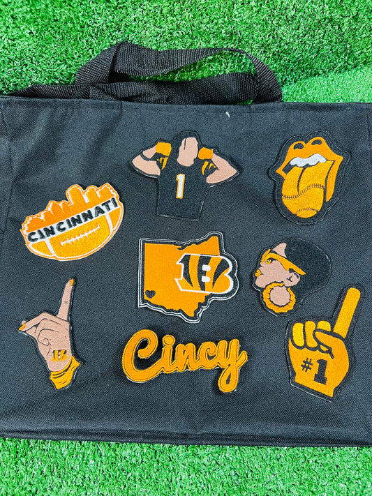 A black tote bag laid flat on green artificial turf, decorated with multiple Cincinnati Bengals-themed embroidered patches including a football, jersey, tongue-and-glossy-stone, map outline, hand gesture, and the word 'Cincy'.