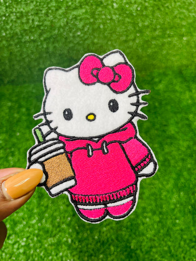 A set of four embroidered patches displayed on bright green grass. The set includes a white kitty wearing a pink hoodie, and holding an iced coffee, a pink bow, a green flower with a yellow center, and a pink flower with a yellow center. The patches are digitized and embroidered with high-quality stitching.