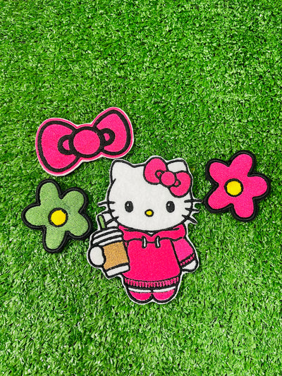 A set of four embroidered patches displayed on bright green grass. The set includes a white kitty wearing a pink hoodie, and holding an iced coffee, a pink bow, a green flower with a yellow center, and a pink flower with a yellow center. The patches are digitized and embroidered with high-quality stitching.