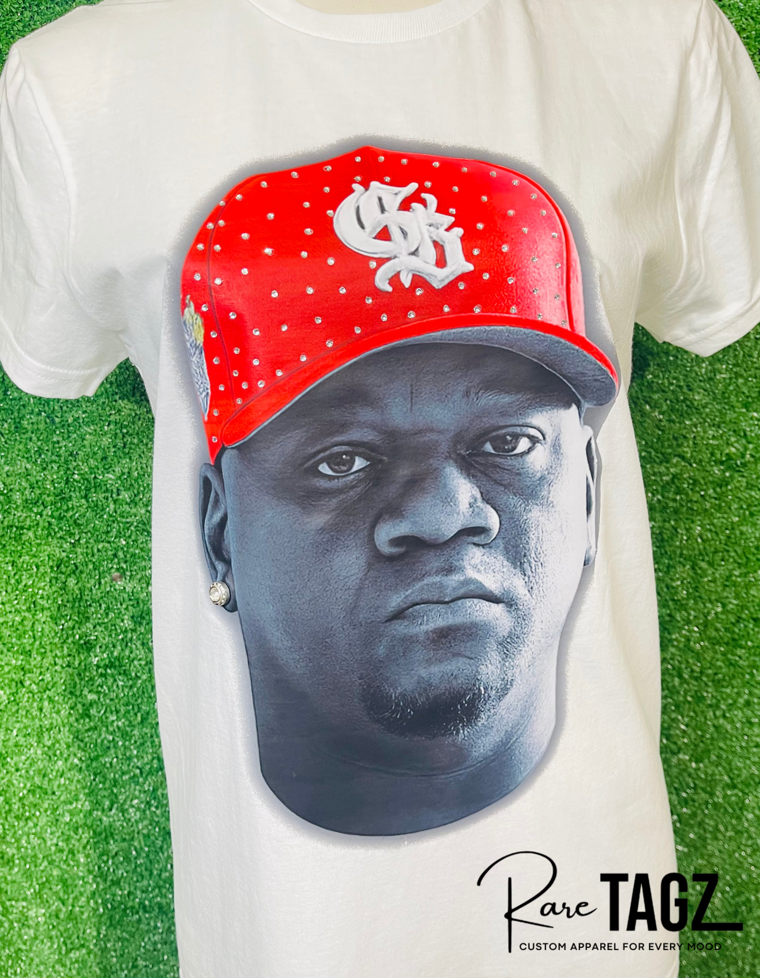 White t-shirt with a graphic of a face wearing a red cap on a grass background, featuring the brand 'Rare TAGZ'.
Custom DJ Fresh portrait t-shirt with hand placed rhinestones on red cap. Custom Breezy Tour shirt by Rare Tagz.