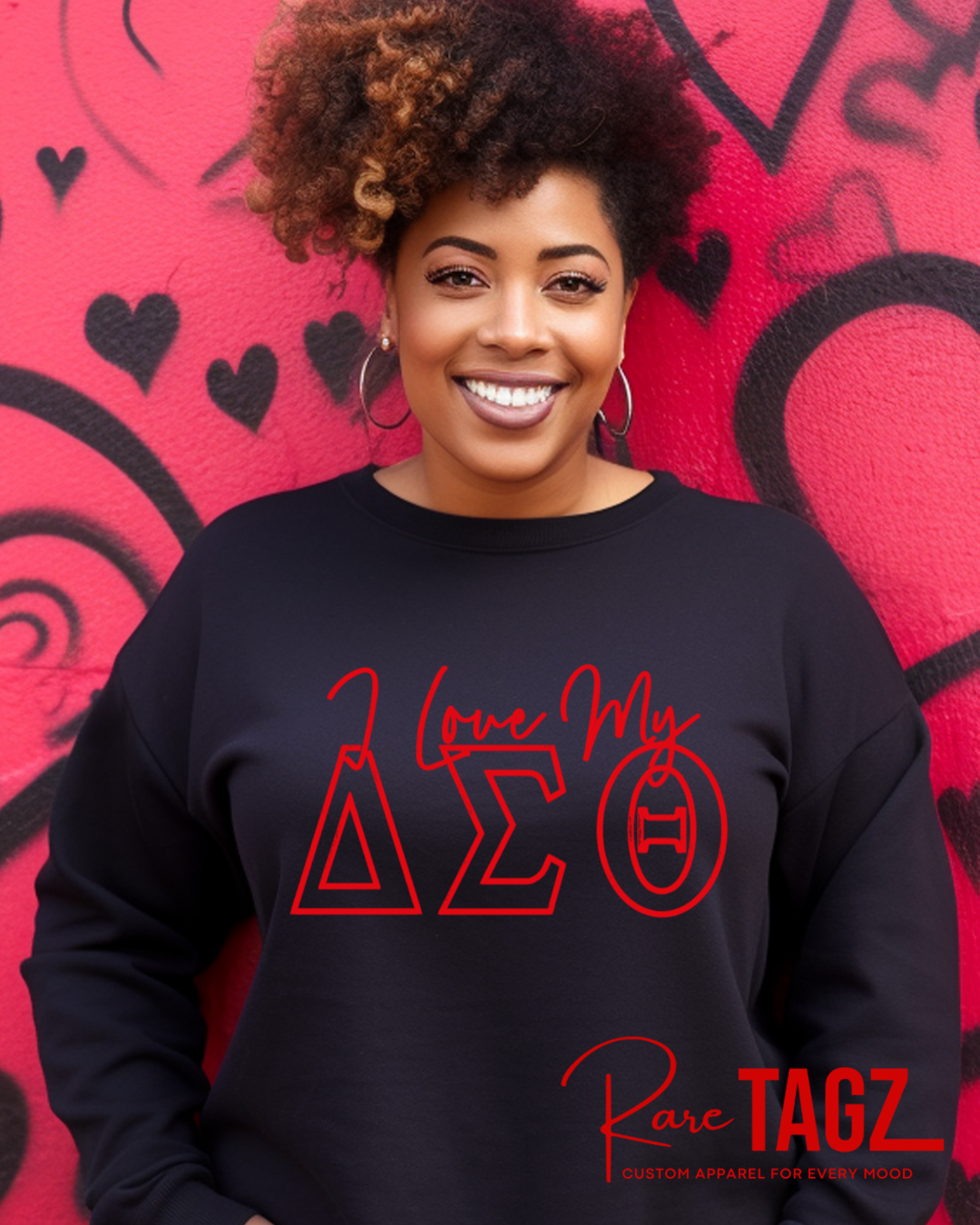 Black Delta Sigma Theta sweatshirt with bold red Greek letters ‘I Love My ΔΣΘ,’ modeled in front of a pink heart graffiti wall. Custom DST paraphernalia by Rare Tagz.