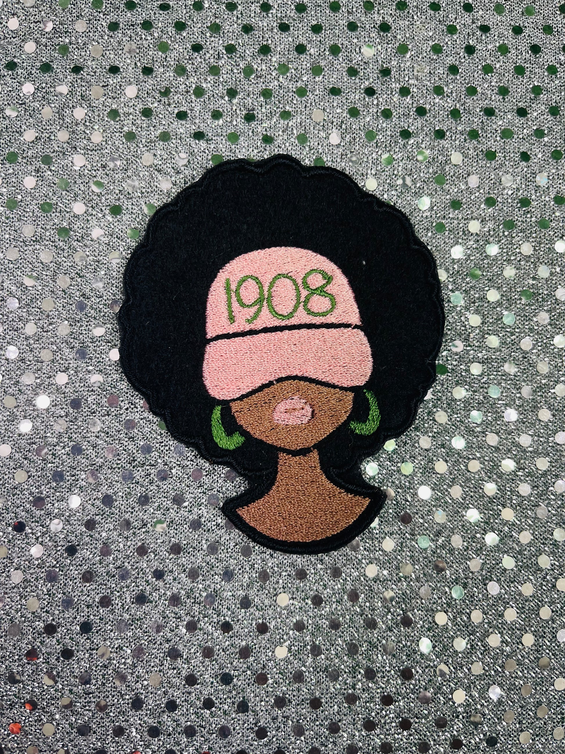 Patch of a woman with an afro, wearing a pink headscarf with '1908' text, on a textured gray background. Alpha Kappa Alpha