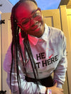 A smiling woman wearing a gray embroidered sweatshirt with the text HE LOVES IT HERE in black featuring a red heart.