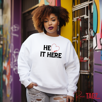 A smiling woman wearing a white embroidered sweatshirt with the text HE LOVES IT HERE in black featuring a red heart.