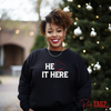 A smiling woman wearing a black embroidered sweatshirt with the text HE LOVES IT HERE featuring a red heart.