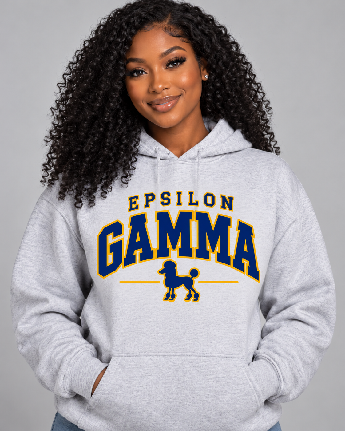 A woman with curly black hair wearing a light gray heather hoodie with 'EPSILON GAMMA' and a poodle graphic printed in navy blue and gold on the front, set against a solid gray studio background.