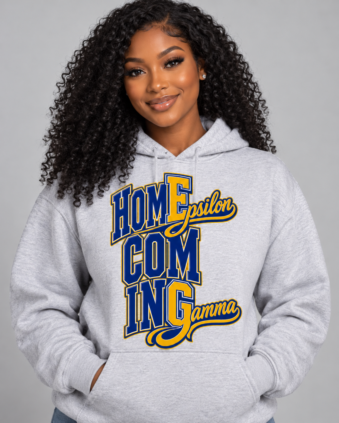 A woman with curly black hair wearing a light gray heather hoodie with a 'Homecoming Gamma' graphic in blue and yellow on the front, set against a solid gray studio background.