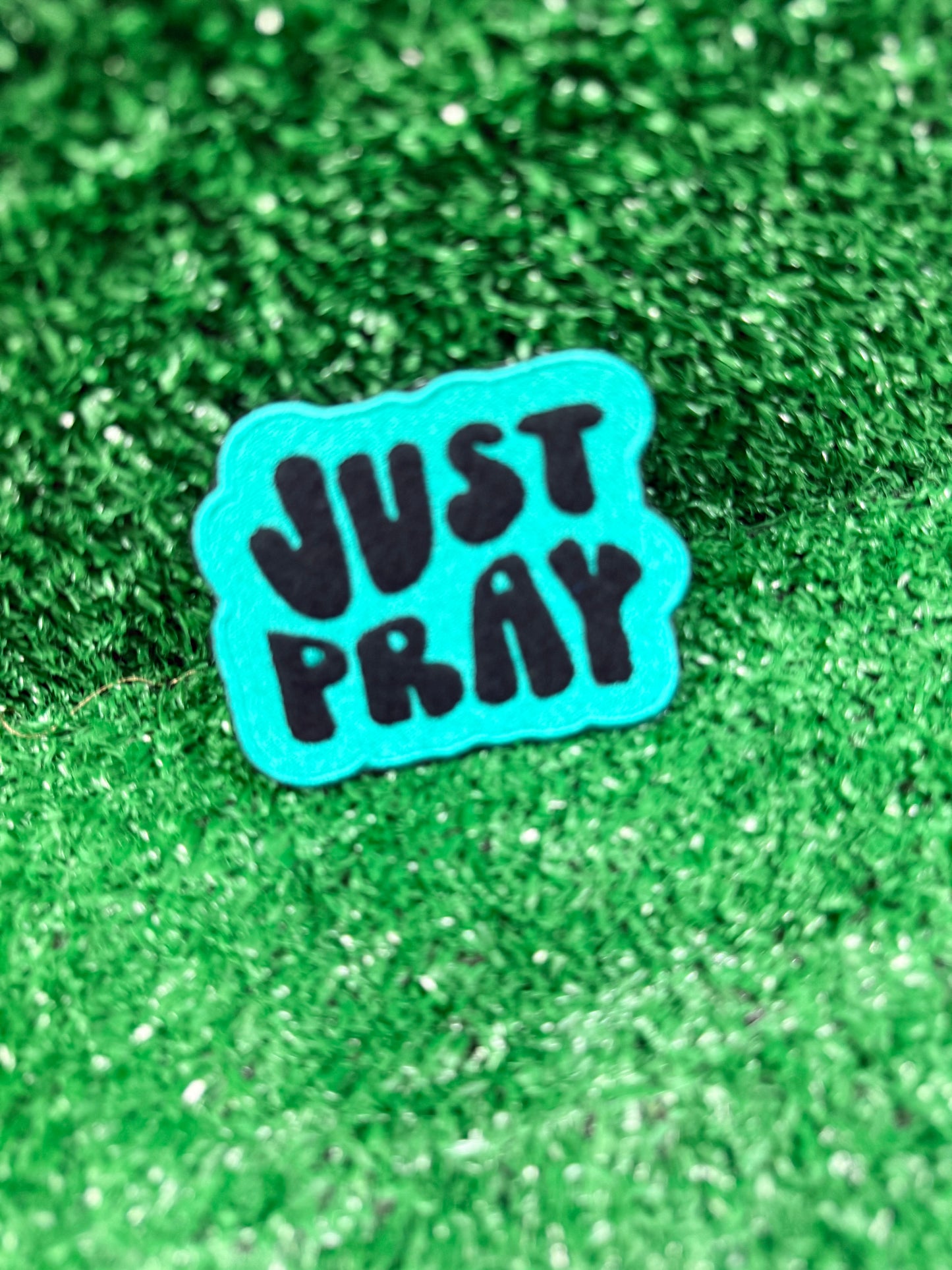 Teal and black 'JUST PRAY' embroidered iron on patch on a green glittery background. Perfect for jackets, hats, backpacks, and more.