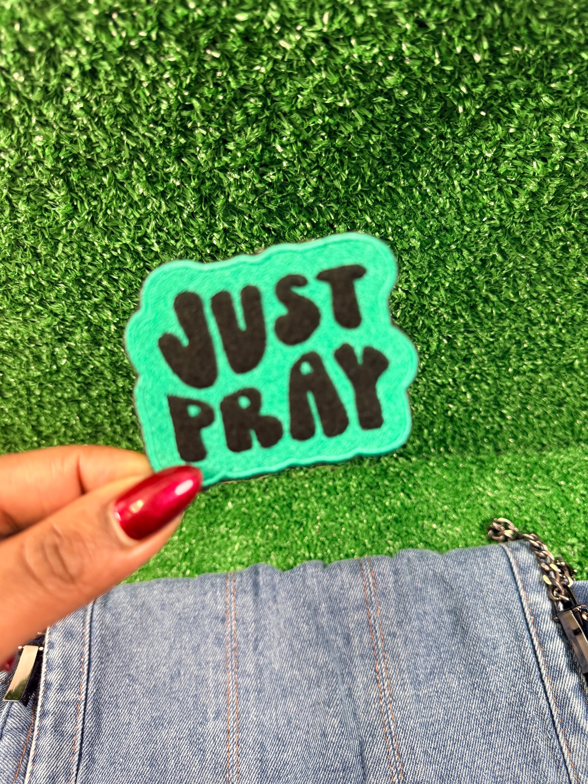 Teal embroidered iron-on patch with 'JUST PRAY' text held over green grass