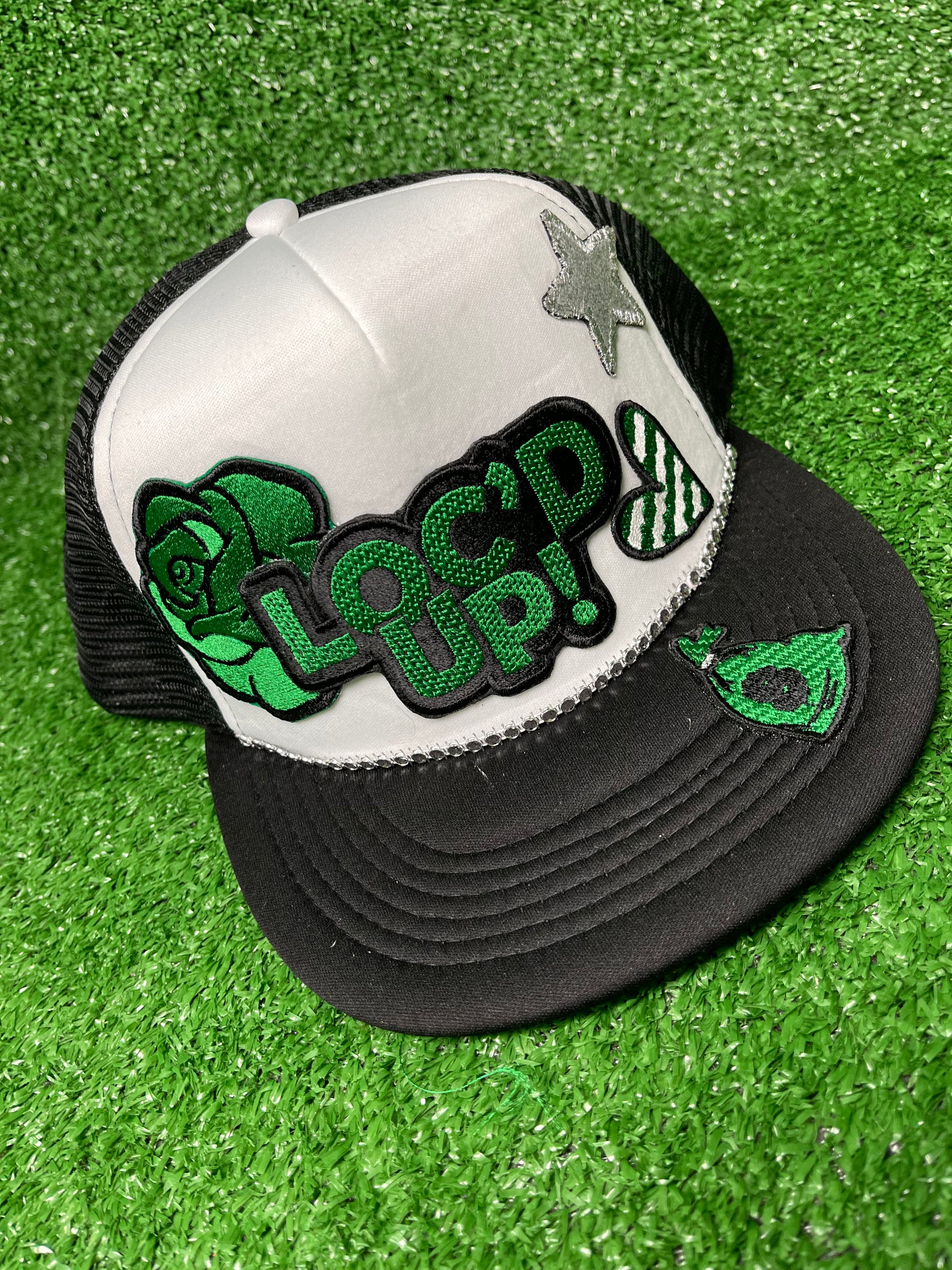 White & Black trucker hat with the text 'Loc’d Up' embroidered in bold black and green letters with other accent patches. The hat features a classic mesh back and adjustable snap closure, perfect for showcasing your locs with style.