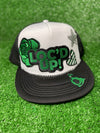 White & Black trucker hat with the text 'Loc’d Up' embroidered in bold black and green letters with other accent patches. The hat features a classic mesh back and adjustable snap closure, perfect for showcasing your locs with style.