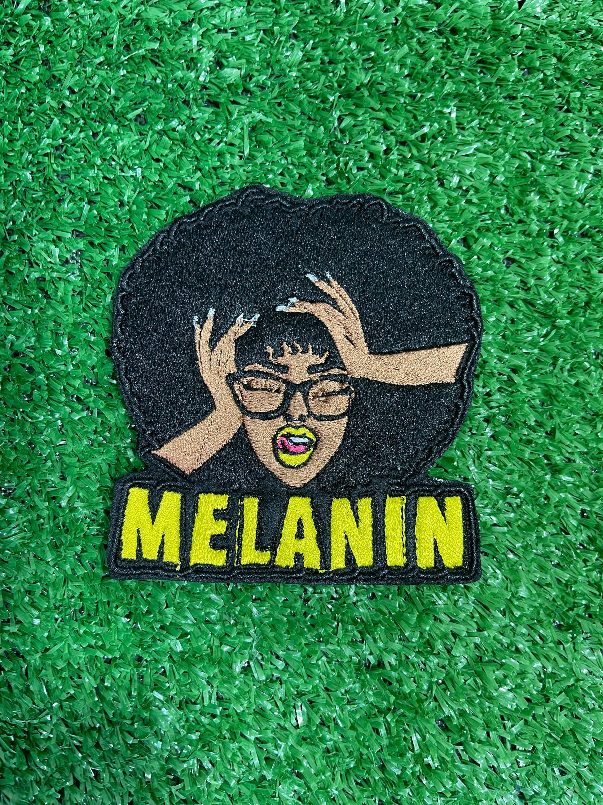 An embroidered patch featuring the word 'MELANIN' and a stylized illustration of a female figure with melonin-rich skin, wearing glasses, and posing with her hands raised.