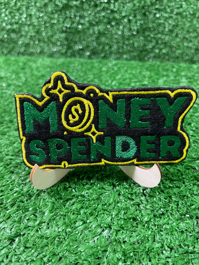 Embroidered iron-on patch reading Money Spender in bold stylish lettering. The patch showcases clean stitching and vibrant green and yellow colors, perfect for customizing clothing or trucker hats.