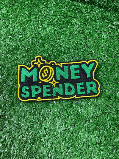 Embroidered iron-on patch reading Money Spender in bold stylish lettering. The patch showcases clean stitching and vibrant green and yellow colors, perfect for customizing clothing or trucker hats.