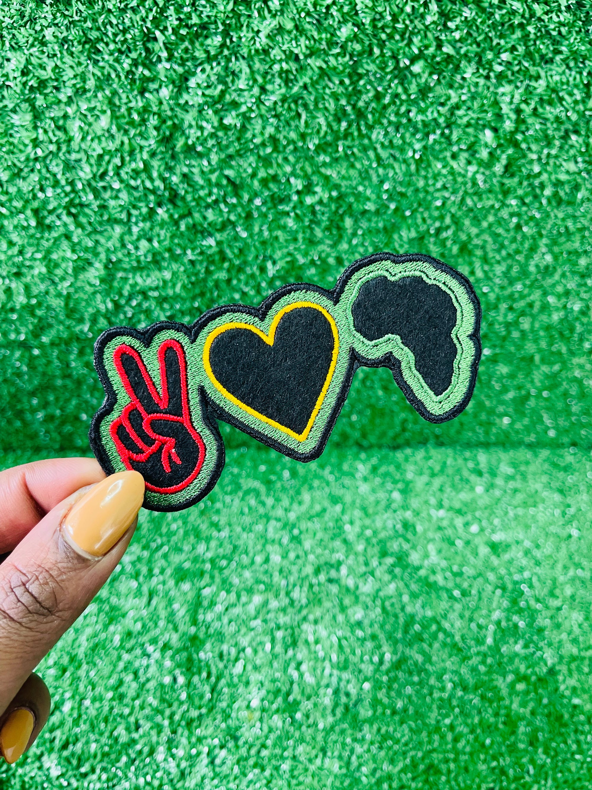 A colorful, embroidered patch featuring a red peace sign a yellow heart, and the silhouette of Africa in green and black. The design symbolizes unity, love, and pride in African heritage.