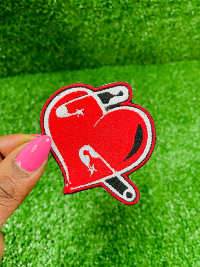 A red embroidered heart-shaped patch with black and white details featuring safety pins piercing through it. The patch has a bold black outline and a slightly distressed look, symbolizing resilience and healing.