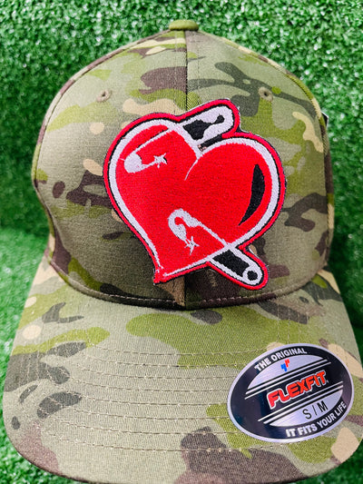 A red embroidered heart-shaped patch with black and white details featuring safety pins piercing through it. The patch has a bold black outline and a slightly distressed look, symbolizing resilience and healing.