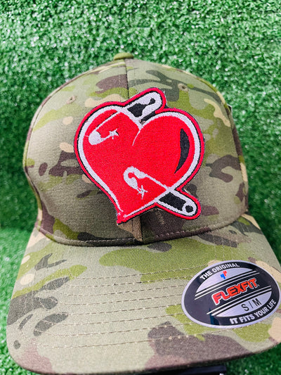 A red embroidered heart-shaped patch with black and white details featuring safety pins piercing through it. The patch has a bold black outline and a slightly distressed look, symbolizing resilience and healing.