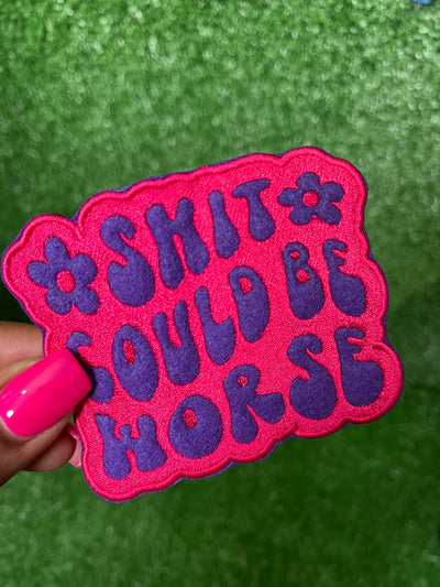 Embroidered patch with the phrase Shit Could be Worse in bold purple lettering on a pink background. The design is simple yet impactful, adding a touch of humor and resilience to any jacket, backpack or trucker hat.