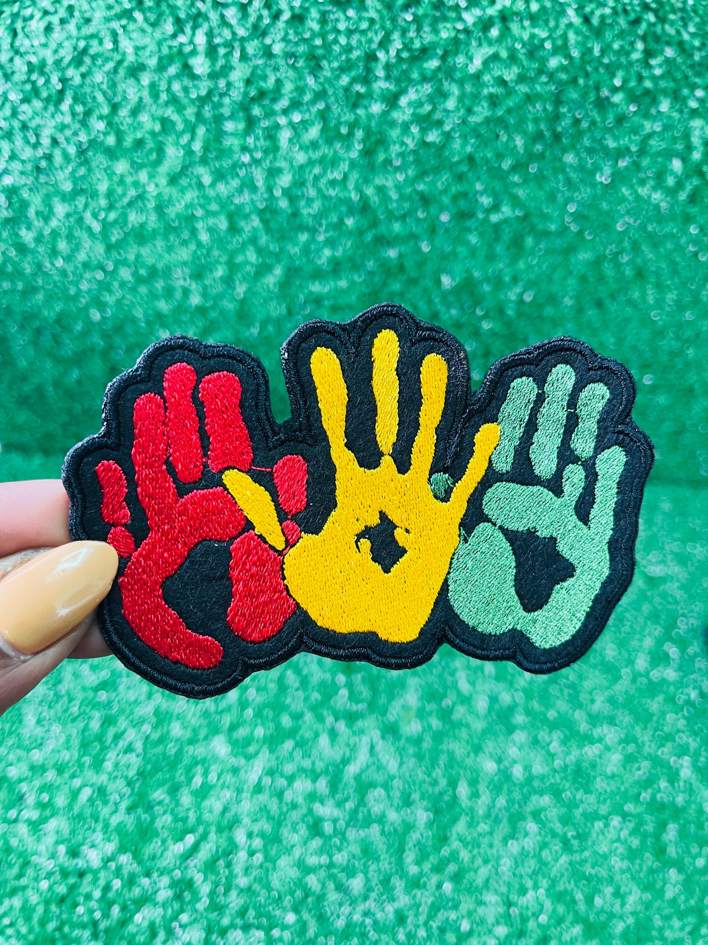 A vibrant embroidered patch featuring three overlapping handprints in red, yellow, green, symbolizing unity. pride, and heritage. The patch has a bold black outline that enhances the striking design. Perfect for celebrating Black culture and adding a meaningful touch to clothing or accessories. 