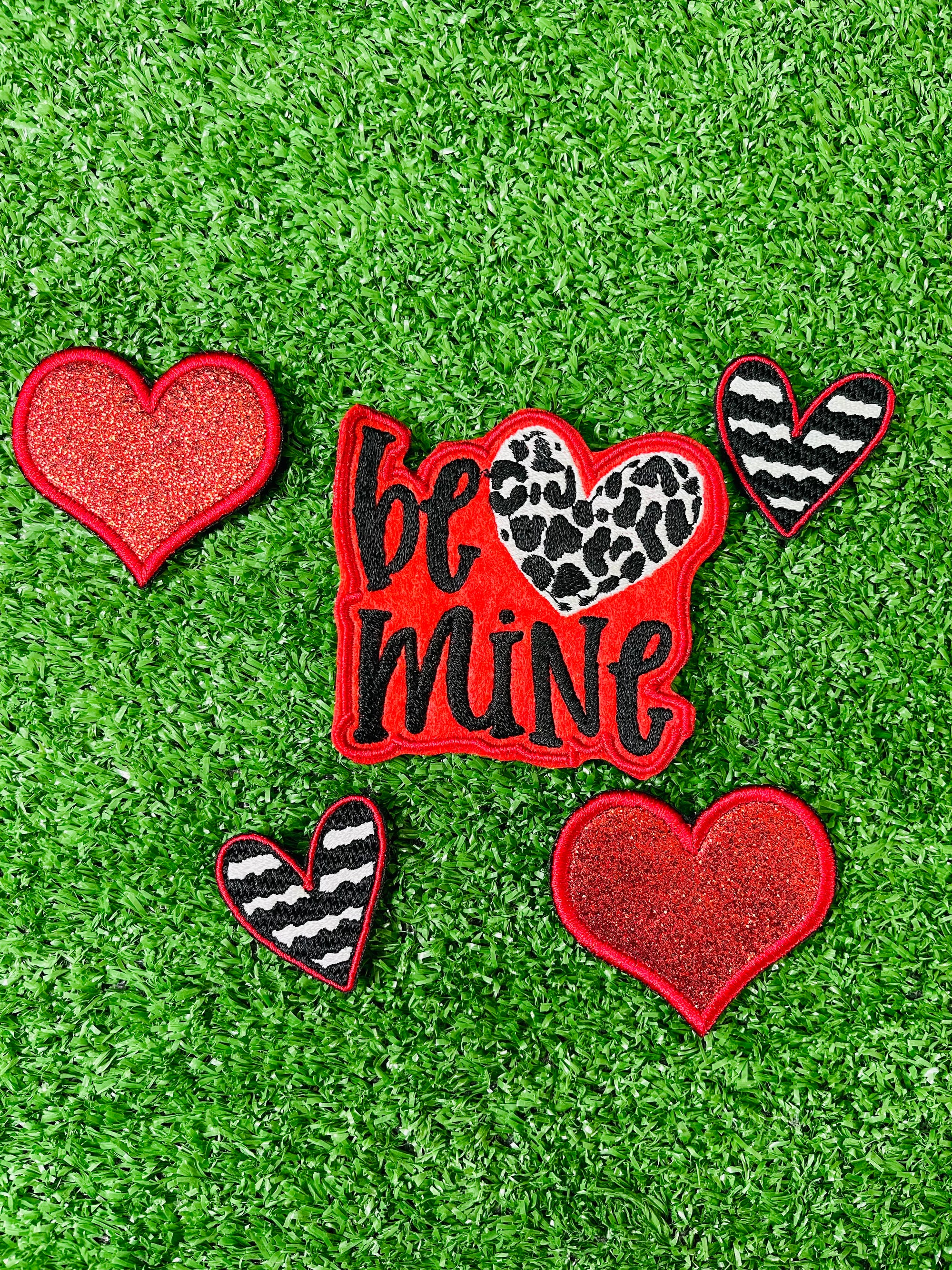 Valentine's Day patch set featuring a bold Be Mine design with a leopard heart, two red glitter hearts, and two black and white zebra stripped hearts displayed on a green grass background.