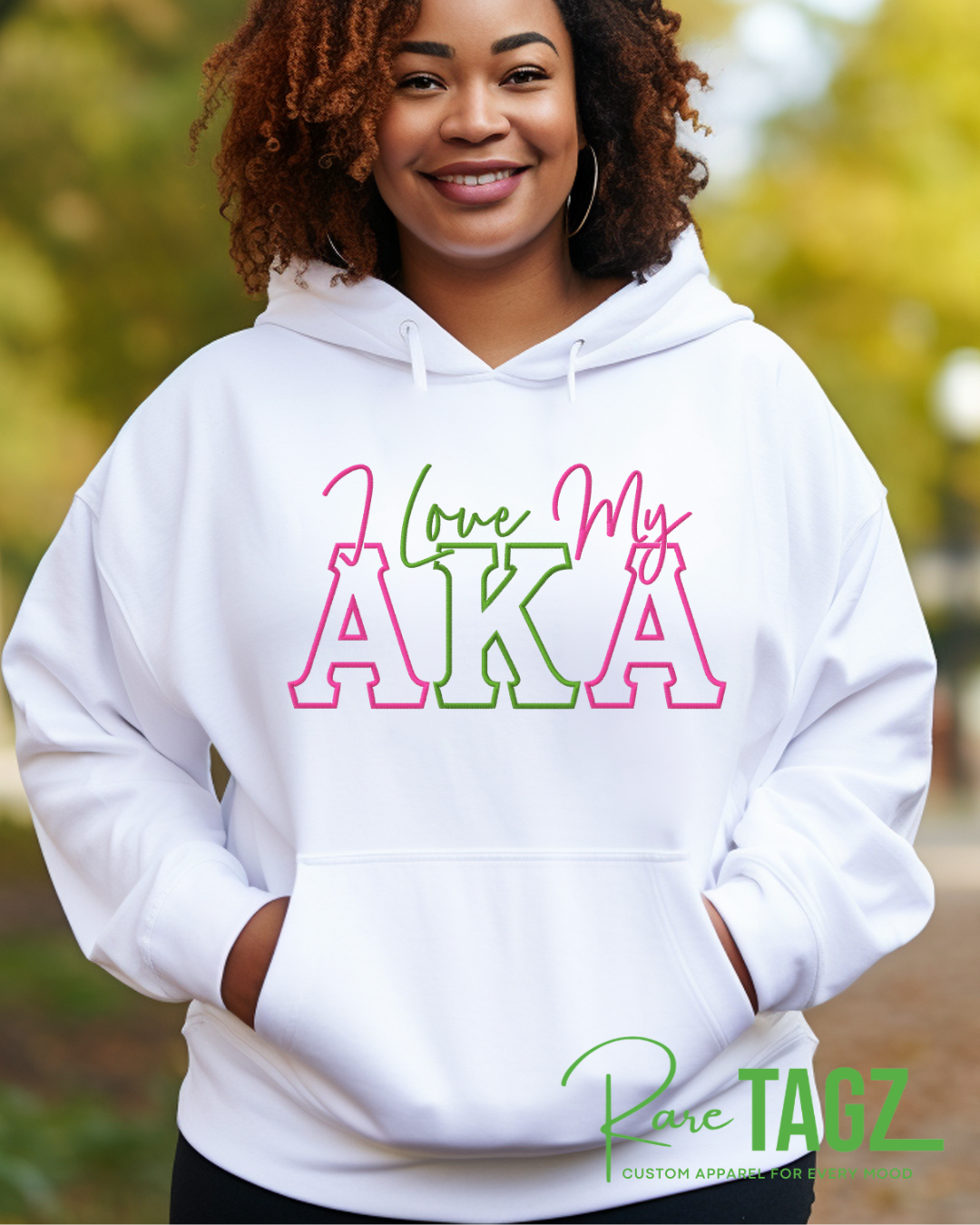 Woman wearing a WHITE hoodie with pink and green lettering that reads ‘I Love My AKA.’ The hoodie has a relaxed fit with a kangaroo pocket and drawstring hood. Background shows colorful graffiti art. Rare Tagz logo is in the corner.”