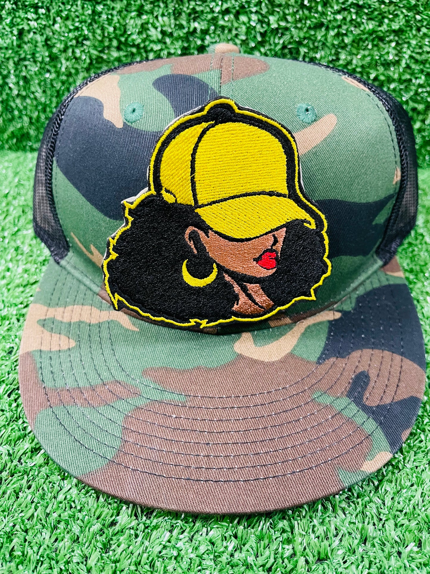 Camouflage cap with embroidered patch of a person wearing a yellow cap on a grass background