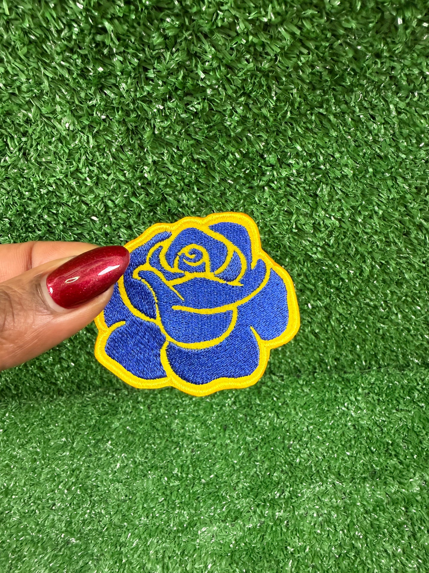 Blue rose embroidered iron onpatch held by a hand on green artificial grass. Perfect for jackets, backpacks, and blazers.