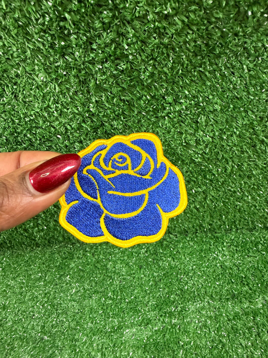 Blue rose embroidered iron onpatch held by a hand on green artificial grass. Perfect for jackets, backpacks, and blazers.